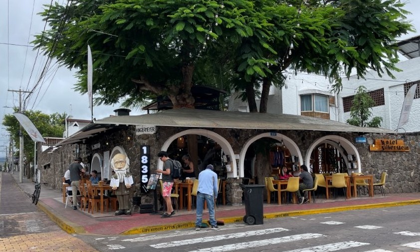 A corner café with white archways and outdoor wooden tables sits beneath a large leafy tree in Puerto Ayora while travelers relax with coffee along the sidewalk. Stopping for coffee at 1835 Café is one of the simple budget-friendly experiences featured in this guide to free things to do on Santa Cruz Island.