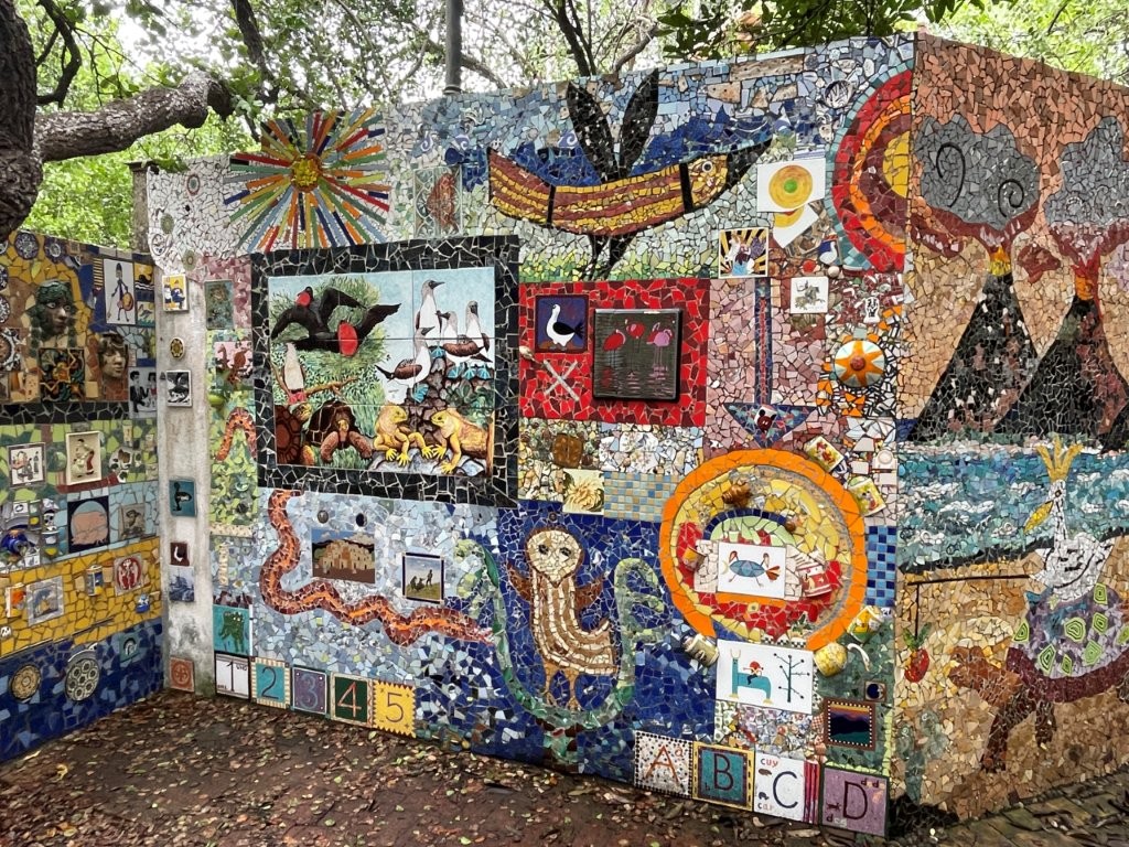 A colorful mosaic wall filled with birds, animals, abstract patterns, and handmade ceramic artwork stands beneath shady trees in the Ceramic Garden. The Ceramic Garden is a quick but creative stop near the Darwin Center that adds a fun artistic break to exploring Santa Cruz Island on a budget.