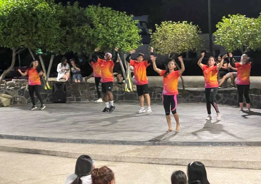 A group of young dancers in matching orange and red shirts perform outdoors at night while visitors gather around the waterfront plaza to watch. Free evening dance performances brought lively local energy to the Puerto Ayora waterfront after dark.