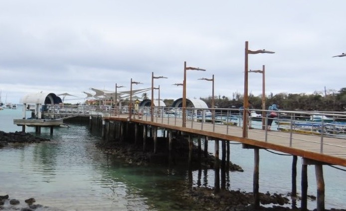 A long wooden ferry pier stretches out over calm turquoise water with boats docked nearby under cloudy skies. Walking the ferry pier and looking for sharks and wildlife below became one of our favorite free things to do on Santa Cruz Island.