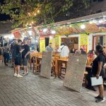 Outdoor restaurants with glowing lights and handwritten menus line a lively pedestrian street filled with diners and travelers at night in Puerto Ayora. Eating along Kiosk Street is a fun and affordable evening activity for travelers looking for cheap local meals on Santa Cruz Island.