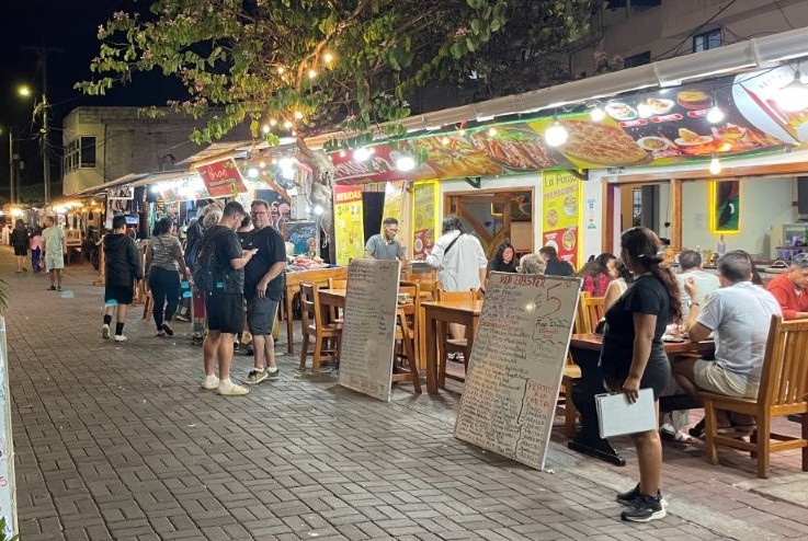 Outdoor restaurants with glowing lights and handwritten menus line a lively pedestrian street filled with diners and travelers at night in Puerto Ayora. Eating along Kiosk Street is a fun and affordable evening activity for travelers looking for cheap local meals on Santa Cruz Island.