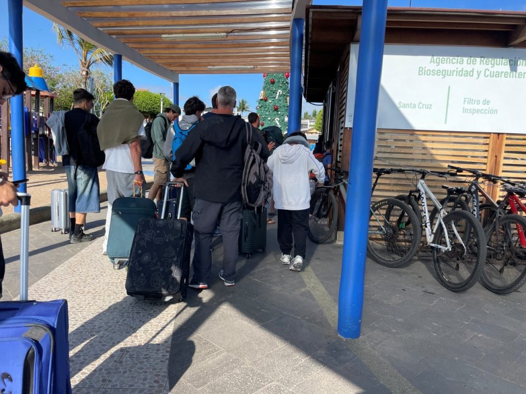 Travelers with rolling suitcases wait outside the Santa Cruz biosecurity inspection area beside blue columns and parked bicycles. Biosecurity screening is part of the Galapagos ferry process, so building in extra time before departure helps the day feel less rushed.