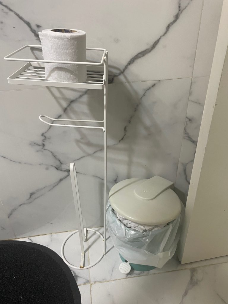 A white metal rack holds a roll of toilet paper while a gray wastebasket sits on the floor below. Toilet paper in the Galapagos Islands needs to be tossed, not flushed.
