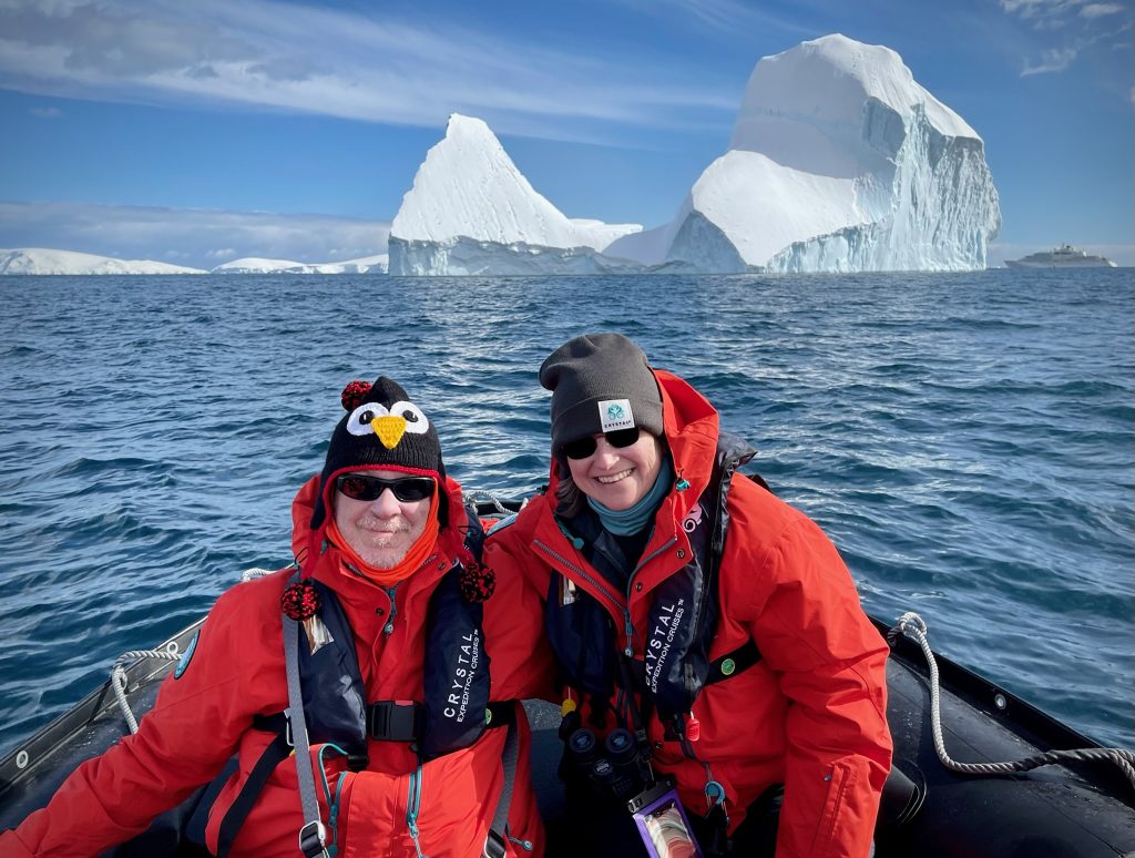 A couple in bright red winter parka jackets sit in the front of the zodiac with large icebergs behind them.