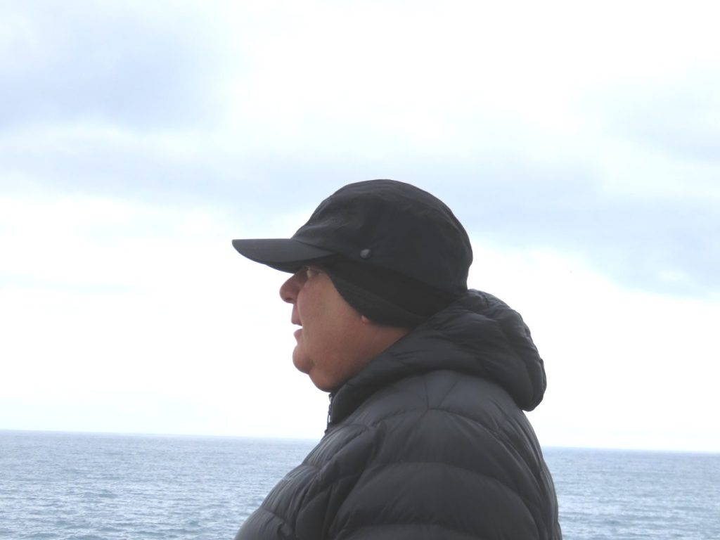 Bill stands on the ship's balcony wearing a black puffer jacket, gloves, knit hat, and athletic shoes. His layering system kept him warm throughout the Antarctica portion of the cruise.
