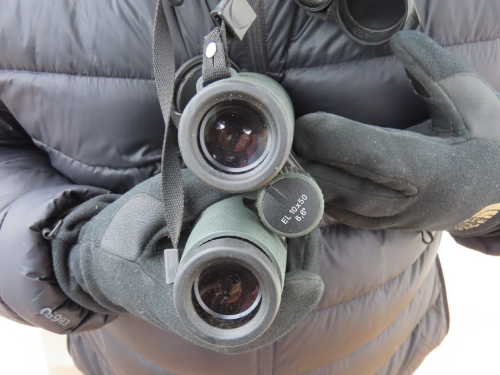 A close-up shows Swarovski binoculars hanging from Bill's neck over a black puffer jacket. Quality binoculars made it much easier to spot whales, seals, penguins, and seabirds before anyone else on deck.