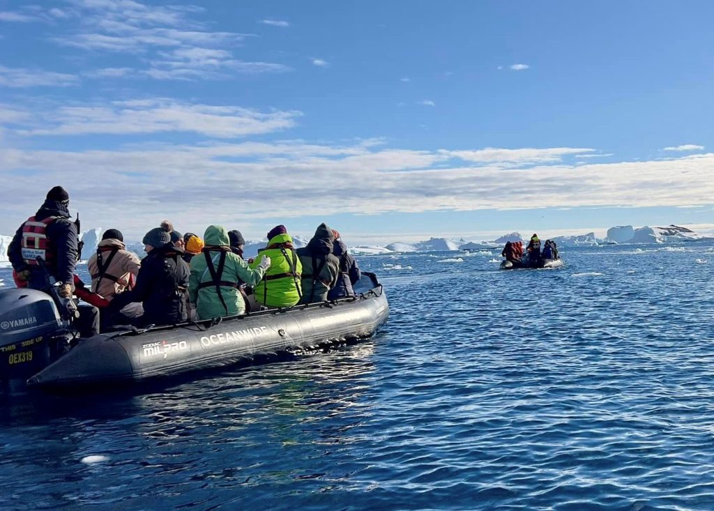 A group of about 12 people ride in a gray zodiac amongst the deep blue waters of Antarctica with ice bergs floating in the distance.