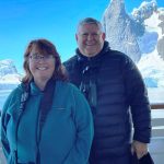 Kari and Bill smile on the deck of their cruise ship with towering Antarctic peaks, glaciers, and floating ice behind them. We almost skipped this Bucket List trip because of the price until we discovered a more affordable option.