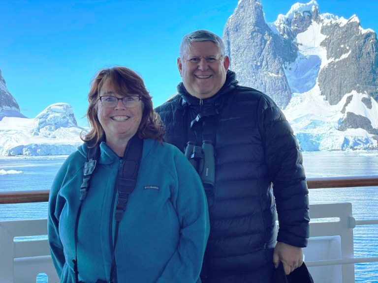 Kari and Bill smile on the deck of their cruise ship with towering Antarctic peaks, glaciers, and floating ice behind them. We almost skipped this Bucket List trip because of the price until we discovered a more affordable option.