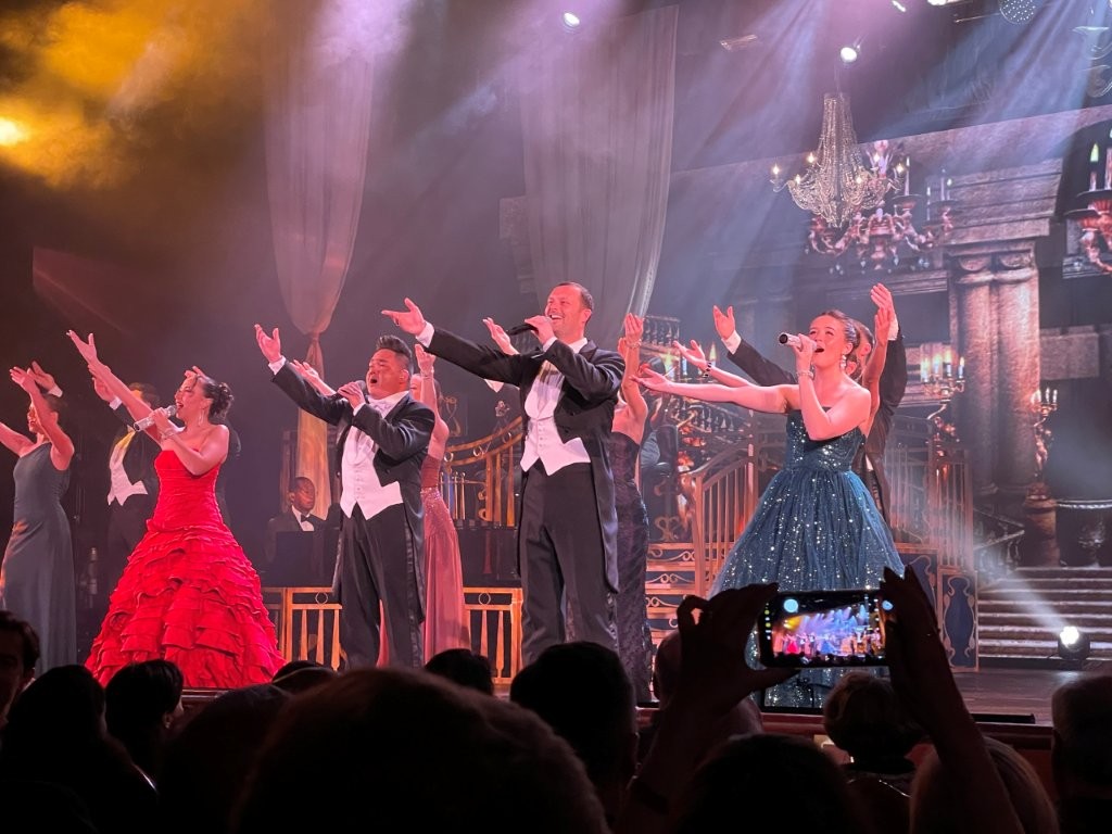 Singers perform on stage beneath large illuminated set pieces while the audience watches from the theater. Most evenings feature a full-scale production show or guest entertainment.