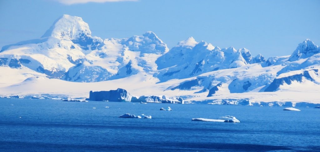 Snowy Antarctic peaks rise above deep blue water dotted with floating ice. 