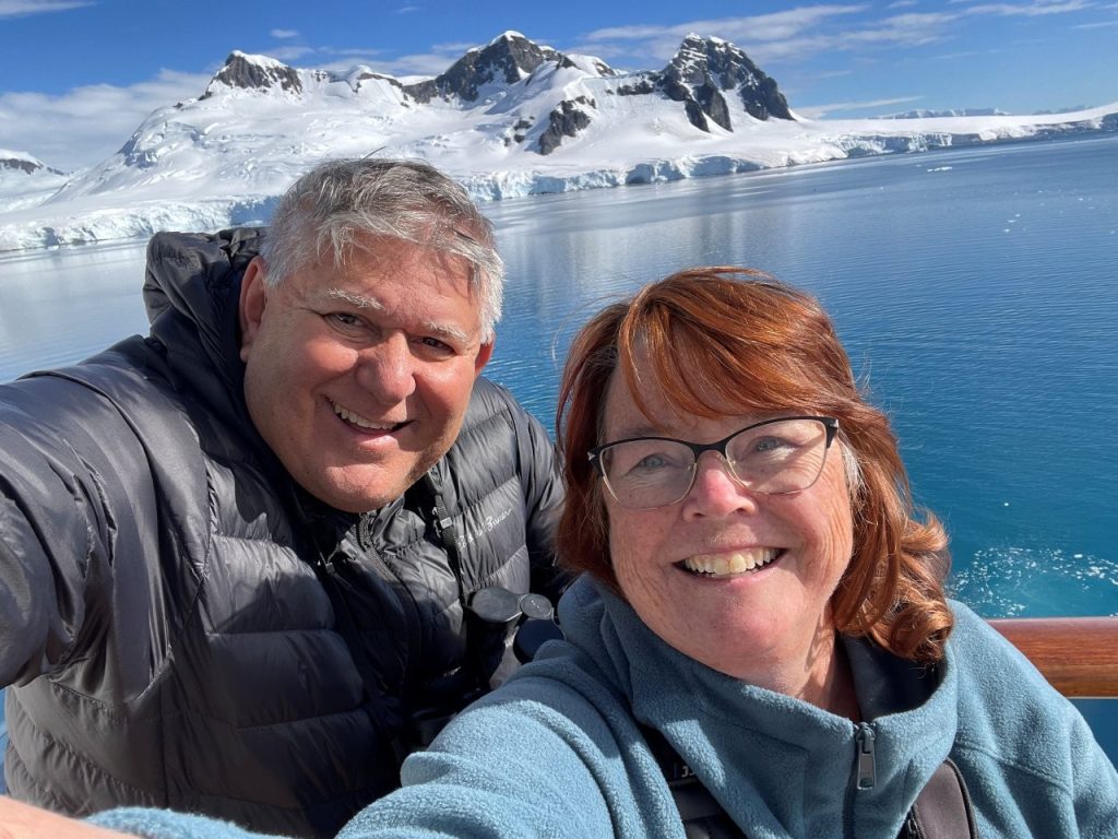 Kari and Bill smile from the cruise ship with snow-covered Antarctic mountains behind them. Antarctica had been at the top of my Bucket List for years, and even Bill was glad we made the journey.