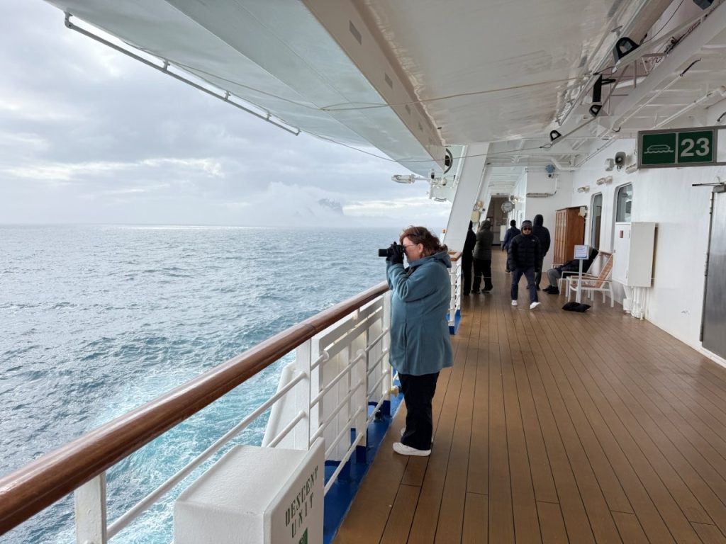 Kari photographs the ocean from the ship while wearing a hooded fleece, gloves, travel pants, and tennis shoes. Layering helped her stay comfortable without packing a bulky parka that she would only wear for a few days.