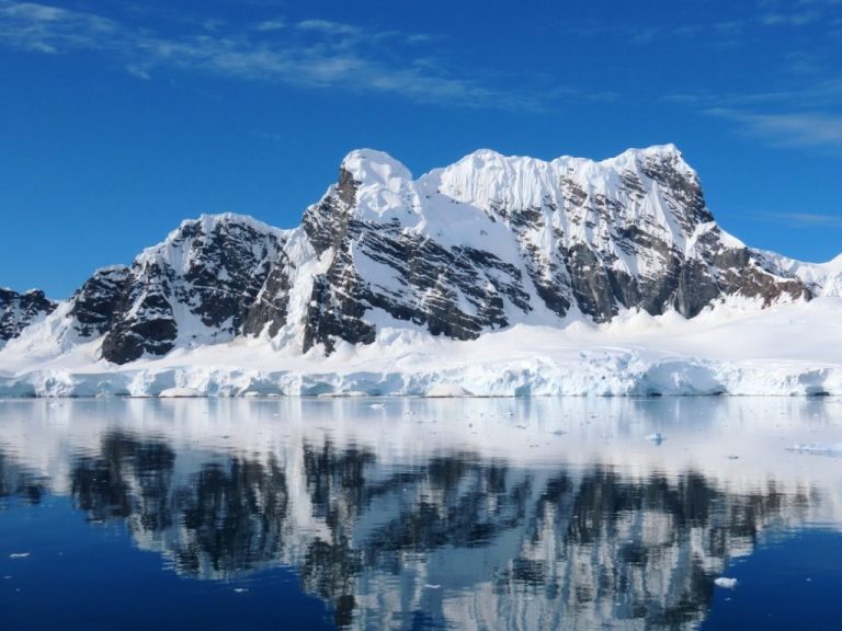 Snow-covered Antarctic mountains reflect perfectly in calm blue water beneath a bright sky. Landscapes like this convinced us our Antarctica cruise was worth every dollar.