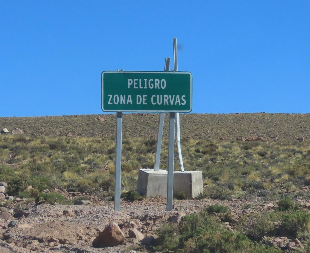 A roadside sign warns drivers of a winding section of road ahead in a remote desert landscape. Paying attention to local traffic signs is especially important when driving through the Atacama's mountainous terrain.