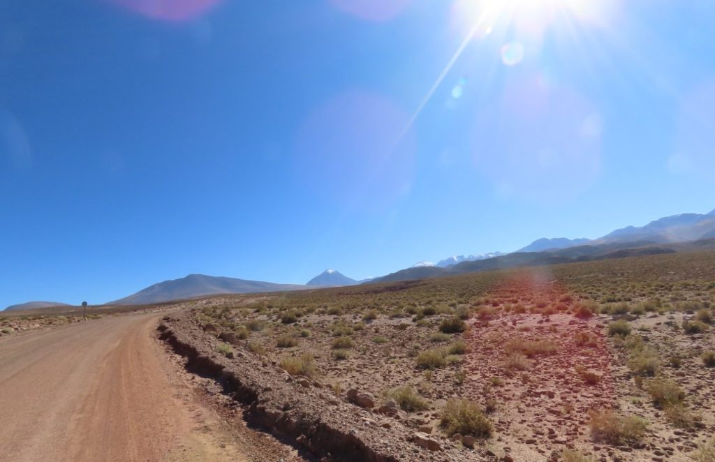 An unpaved road stretches across open desert toward distant snow-dusted volcanoes under intense midday sun. Some sections offered enormous views, while the next turn could close in between hills and rocks.