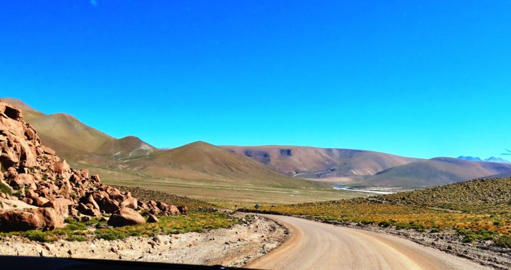 A broad green wetland spreads beneath rounded brown hills and a deep blue sky, with a distant volcano rising between the slopes. The higher Atacama Desert altitude leaves some people lightheaded and tired.