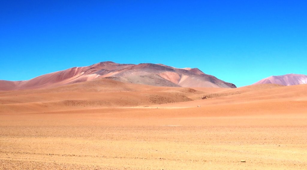 Pale orange desert hills roll toward a distant pink mountain beneath an intense blue sky. This wide-open stretch shows why Ruta 27 felt remote even though the road itself was paved and easy to follow.