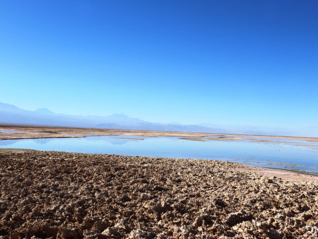 White salt flats and a narrow band of water stretch across a broad red-brown basin below dark volcanic mountains. This roadside view shows the more mineral, remote-feeling landscape near Salar y Laguna de Tara.