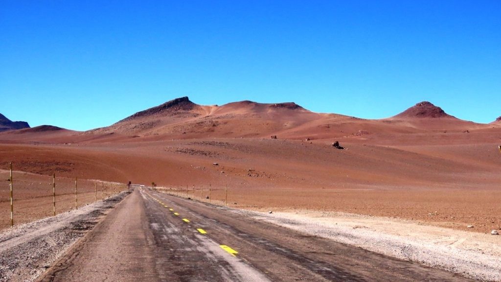 A narrow paved highway disappears into rolling red hills beneath an endless blue sky. One of the joys of visiting the Atacama Desert is simply driving through landscapes that seem to go on forever.