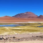 White salt bands curl around blue and pink mineral pools below red volcanic hills and a deep blue sky. This image captures the wild, high-altitude scenery that makes Ruta de los Salares one of the most dramatic drives from San Pedro de Atacama.
