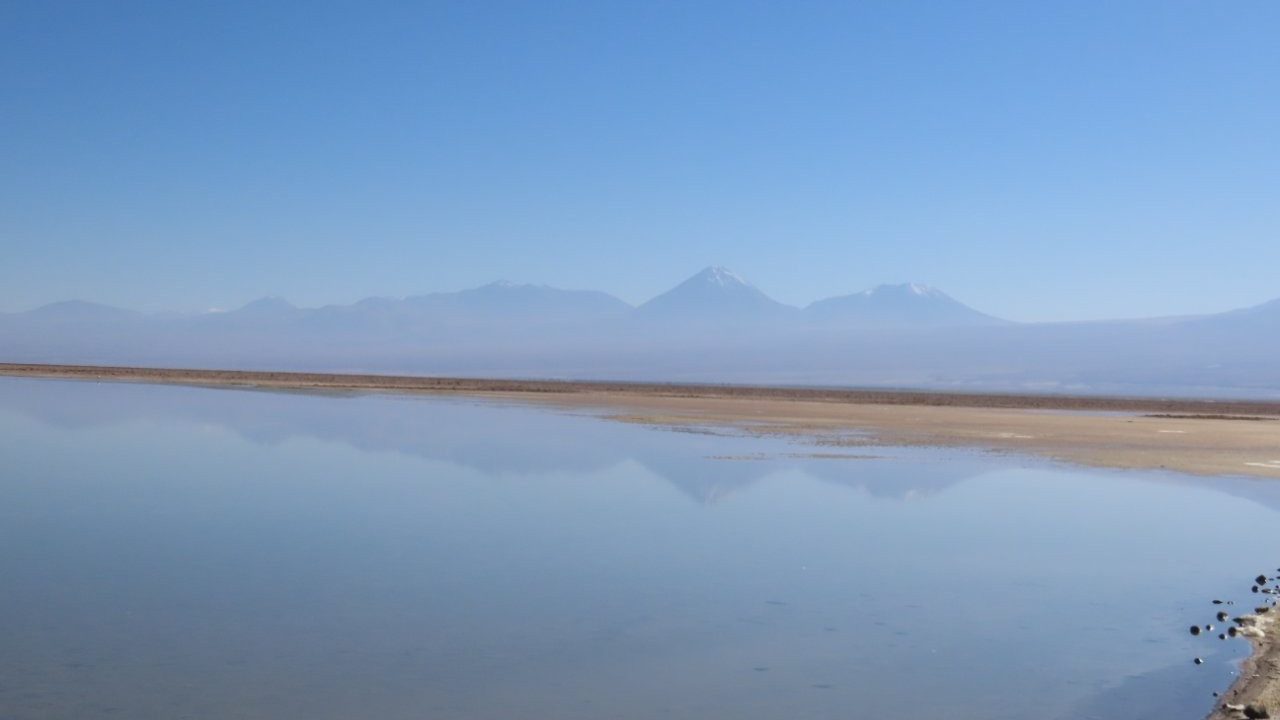 Snow-dusted volcanoes rise beyond a calm lagoon that reflects the mountains and pale blue sky. The peaceful scenery made this lower-altitude drive feel completely different from the busier attractions near San Pedro.