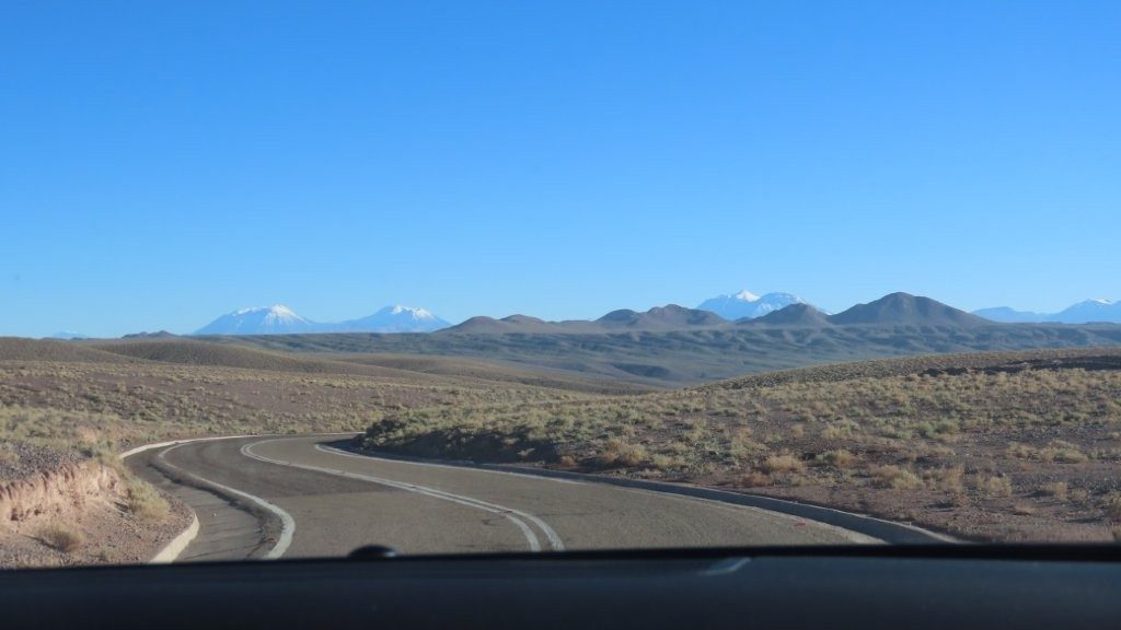 A paved road curves through rolling brown hills toward a line of snow-covered volcanoes on the horizon. The smooth start along B-207 gave little warning that the road ahead would soon become much less obvious.