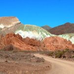 A dusty road curves toward layered red, green, cream, and white mountains beneath a brilliant blue sky. A guided tour allows you to see the unusual colors at Rainbow Valley Atacama.