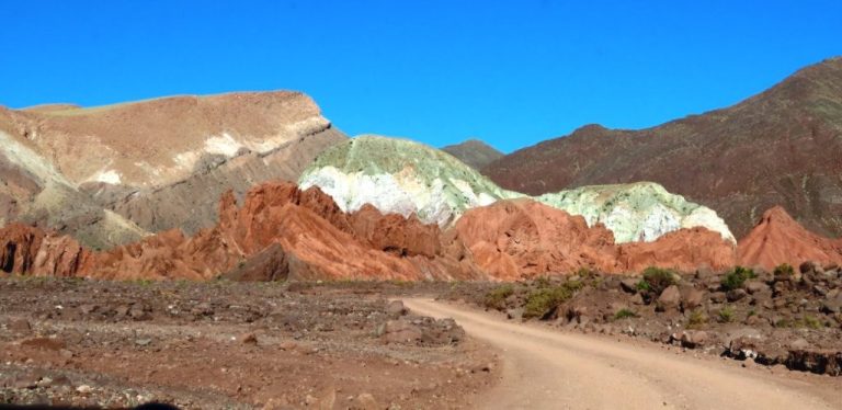 A dusty road curves toward layered red, green, cream, and white mountains beneath a brilliant blue sky. A guided tour allows you to see the unusual colors at Rainbow Valley Atacama.