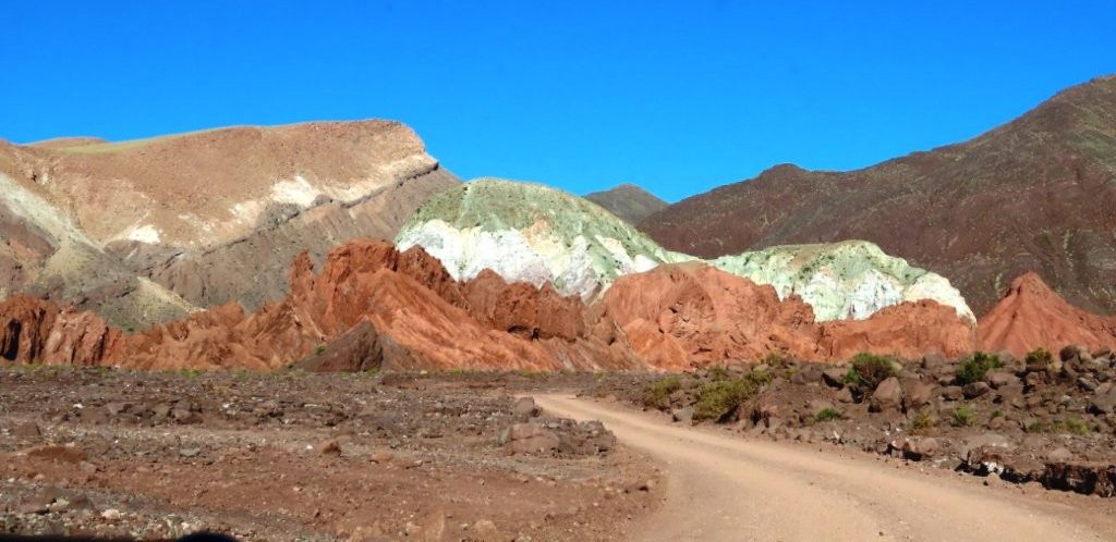 A dusty road curves toward layered red, green, cream, and white mountains beneath a brilliant blue sky. The route captures both the unusual colors and the remote driving experience at Rainbow Valley Atacama.