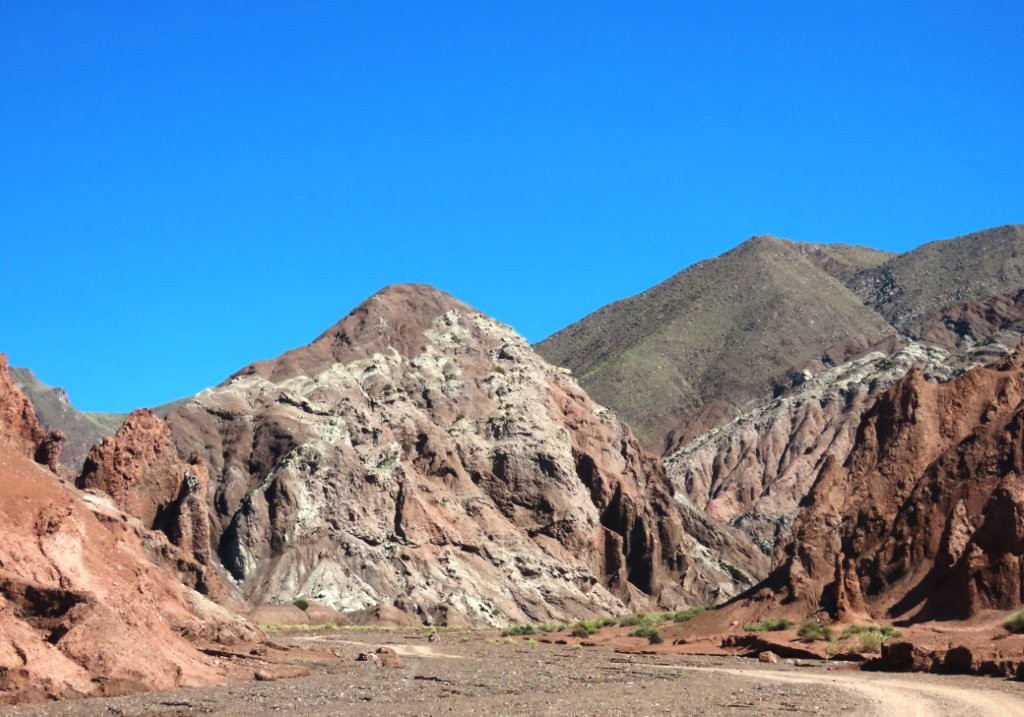A winding dirt road disappears into a canyon surrounded by multicolored mountains. The remote setting makes Rainbow Valley feel far removed from the busier attractions near San Pedro de Atacama.