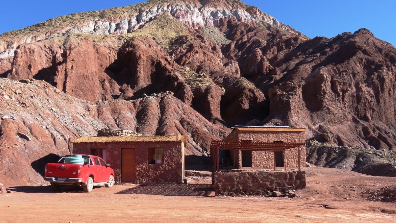 A red pickup truck sits beside two small stone buildings at the base of towering rust-brown cliffs. This is the ticket office for Rainbow Valley