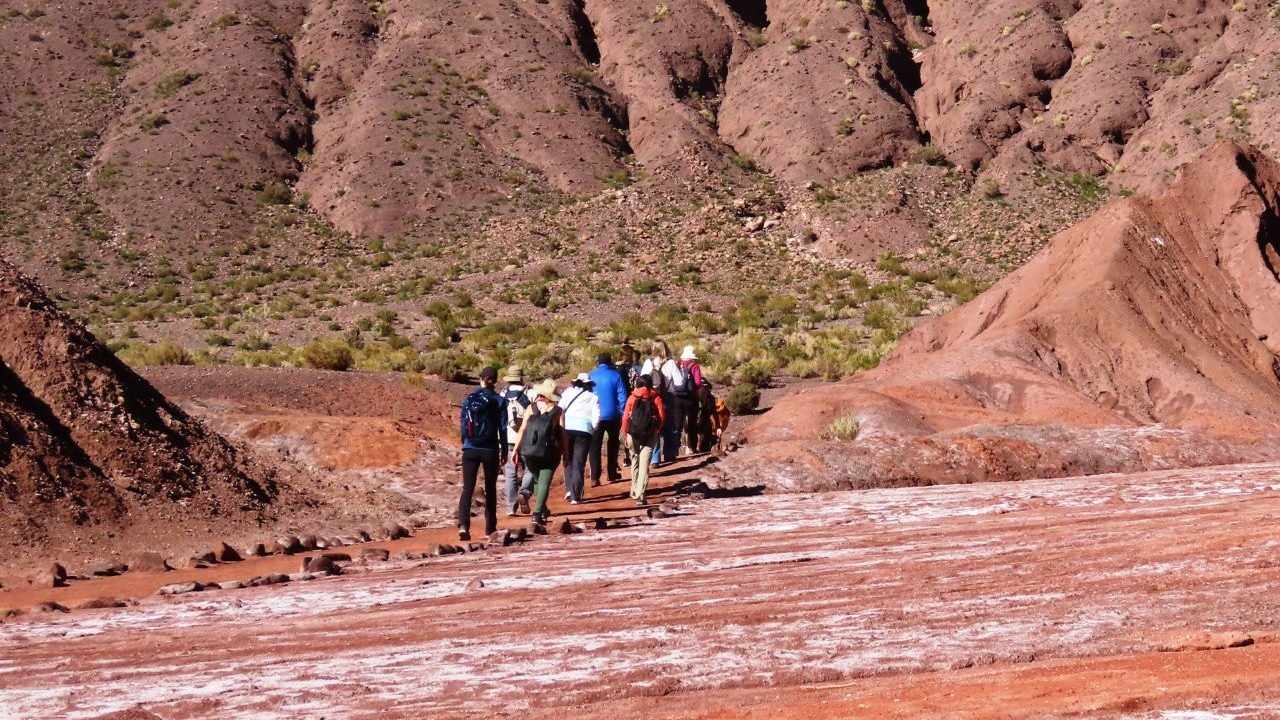 A group of hikers follows a narrow route across red ground streaked with white mineral deposits between steep brown hills of Rainbow Valley. The trail was not clearly marked, so we followed a tour group that seemed to know where it was going.