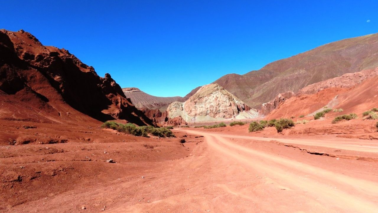 A reddish dirt road bends between dark cliffs and pale mineral-streaked mountains beneath a cloudless sky. Roads around Rainbow Valley Atacama often looked more like flattened tracks than established routes.