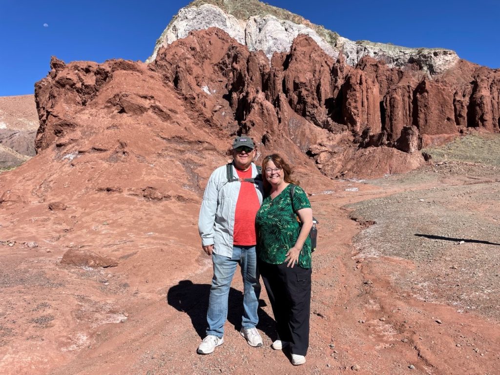 Bill and Kari stand together on red desert ground with jagged rust-colored cliffs and pale mineral bands rising behind them. We were excited to explore the valley, but the 11,000-foot elevation made even the short trail feel harder than expected.