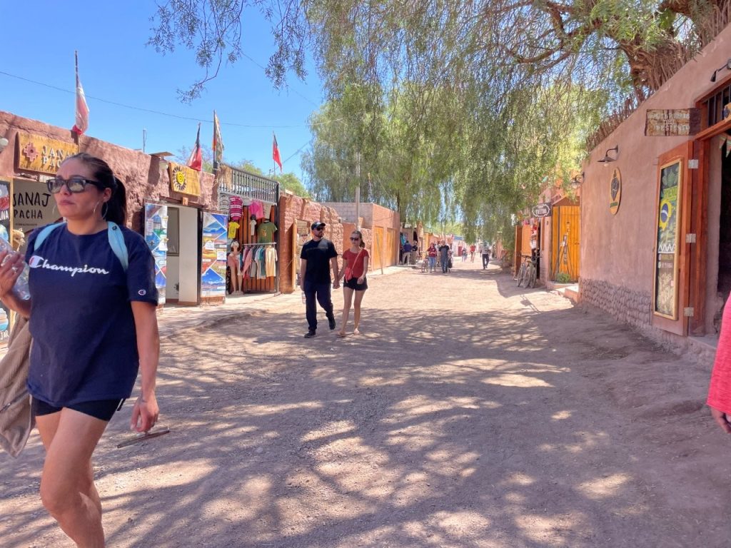 Visitors stroll along a dusty pedestrian street lined with adobe buildings, shops, and shade trees in San Pedro de Atacama. The town's walkable center serves as the main hub for tours, restaurants, and local shopping.