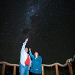 Bill and Kari stand on a wooden viewing platform beneath a dazzling river of stars stretching across the night sky. Stargazing tours take visitors to some of the darkest skies on Earth for an unforgettable evening beneath the Milky Way.