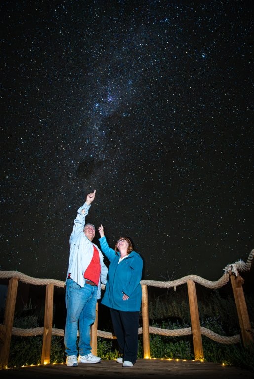 Bill and Kari stand on a wooden viewing platform beneath a dazzling river of stars stretching across the night sky. Stargazing tours take visitors to some of the darkest skies on Earth for an unforgettable evening beneath the Milky Way.