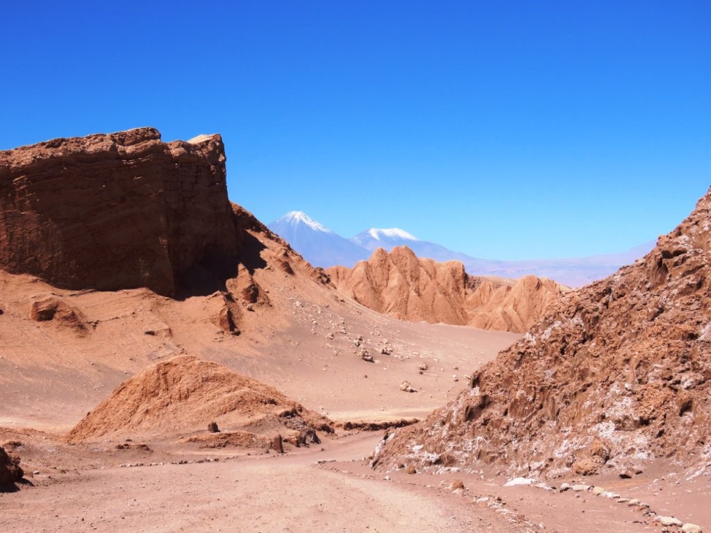 A winding dirt road passes through towering red rock formations with Licancabur Volcano rising in the distance beneath a cloudless blue sky. Scenes like this are why driving through Valle de la Luna is one of the highlights of the Atacama Desert.