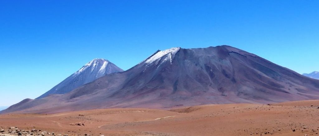 Two snow-dusted volcanoes rise from a barren red plateau under a cloudless blue sky. Many guided tours travel through landscapes framed by the Andes' towering volcanoes.