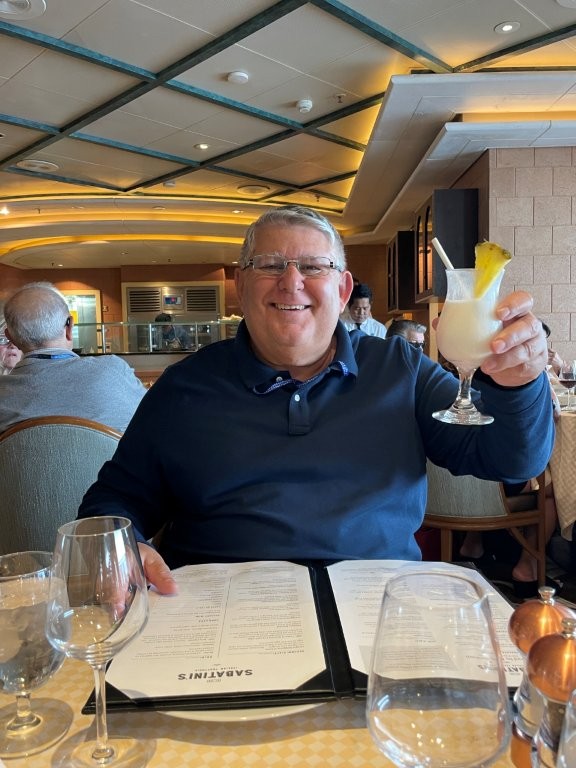 Bill smiles at a cruise ship restaurant table while holding up a creamy tropical cocktail garnished with pineapple. His drink is one example of why a cruise drink package worked well for Bill and Kari on their Antarctica cruise.