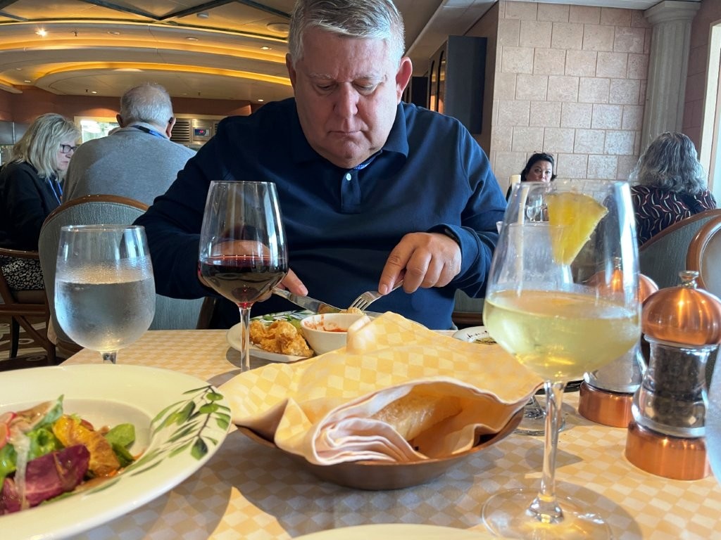 Bill eats lunch at a cruise ship restaurant with glasses of red wine and a pale tropical drink on the table in front of him. With drinks already included in their package, Bill drank more on the Antarctica cruise than he normally does at home.