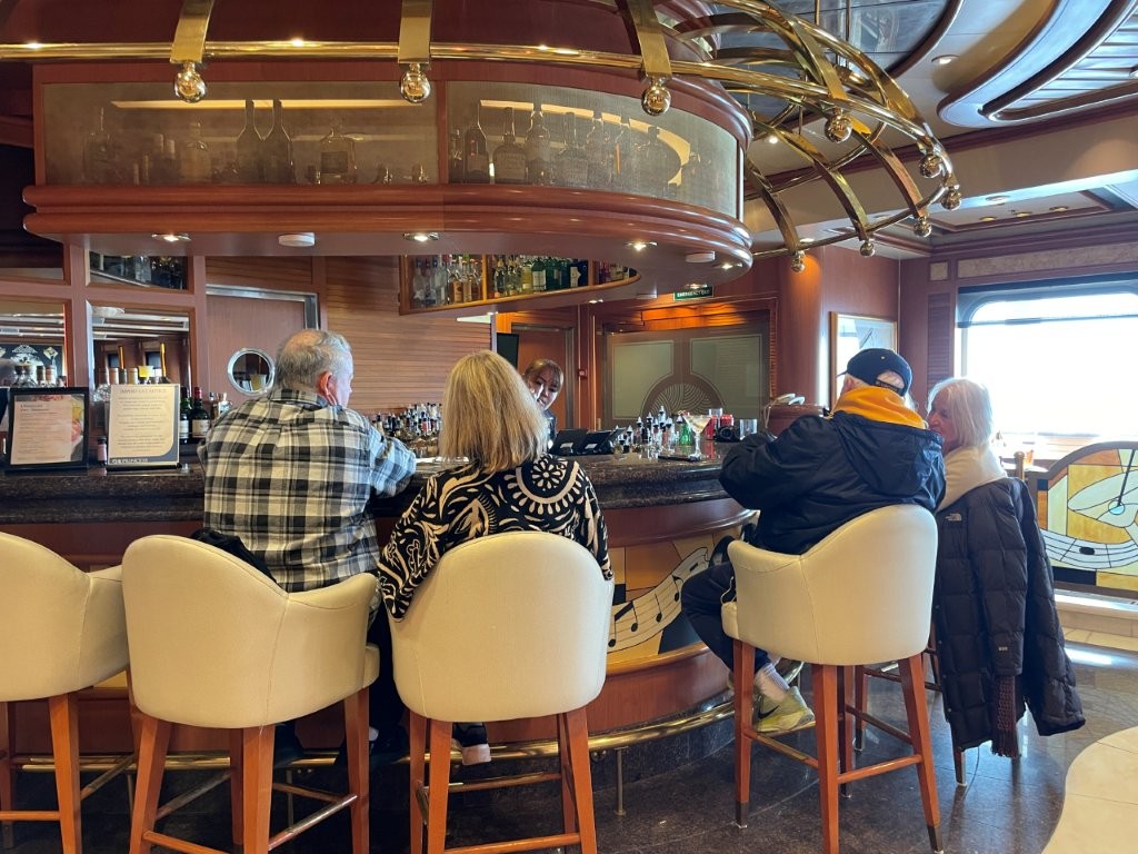 Passengers sit on cream-colored stools around a polished wood cruise ship bar while a bartender serves drinks behind the counter. A cruise drink package makes the most sense when the drinks and other included benefits match what a traveler would normally buy onboard.