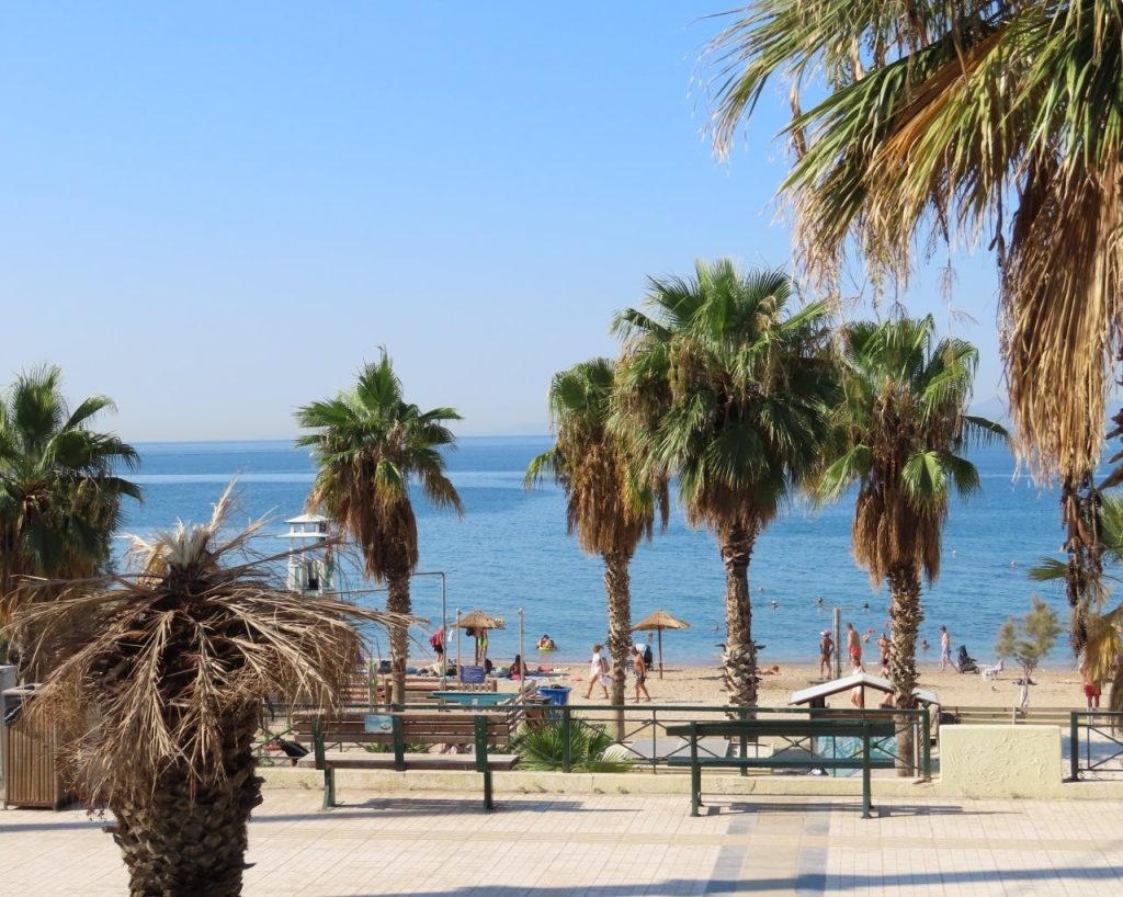 Palm trees frame a sunny stretch of the Athens Riviera with blue Aegean water beyond the waterfront. The coastal scenery was a welcome change once our bus reached the Riviera outside central Athens.