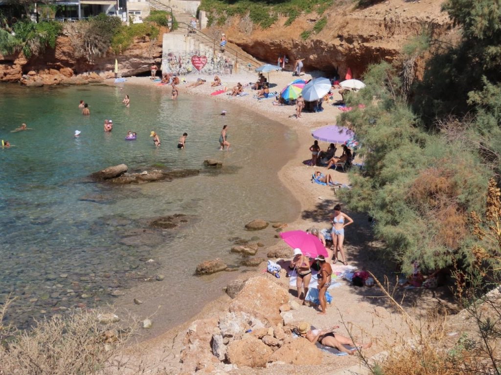 Swimmers float in clear blue water beside the rocky coastline near Athens, Greece, with people gathered along the shore. The rocky entry into the Aegean Sea is the type of beach where sturdy water shoes can make getting in and out of the water easier.