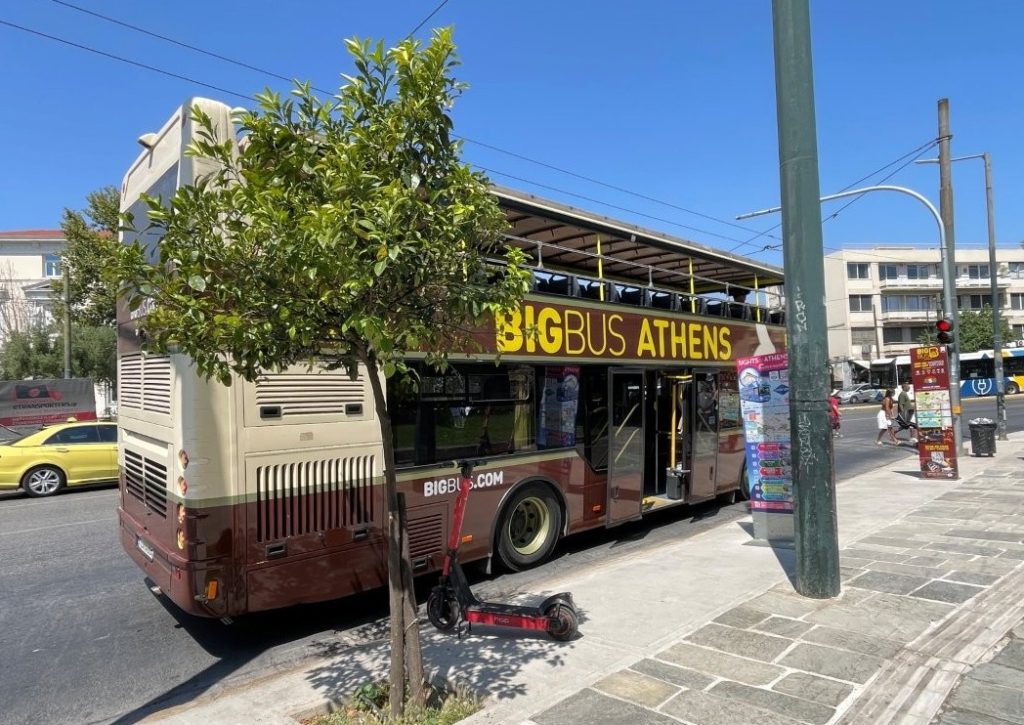A brown sightseeing bus marked “Big Bus Athens” waits along a sunny Athens street beside leafy trees and white buildings. Kari and Bill used the Hop-On/Hop-Off bus for both sightseeing and transportation while exploring Athens.