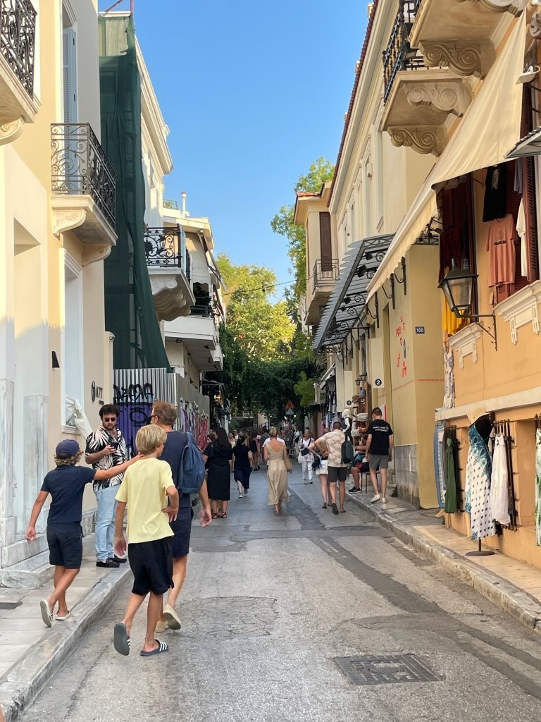 Pedestrians walk along a narrow Plaka street lined with shops, balconies and awnings beneath a bright blue Athens sky. The lively street scene captures one of the central Athens neighborhoods we explored on foot.