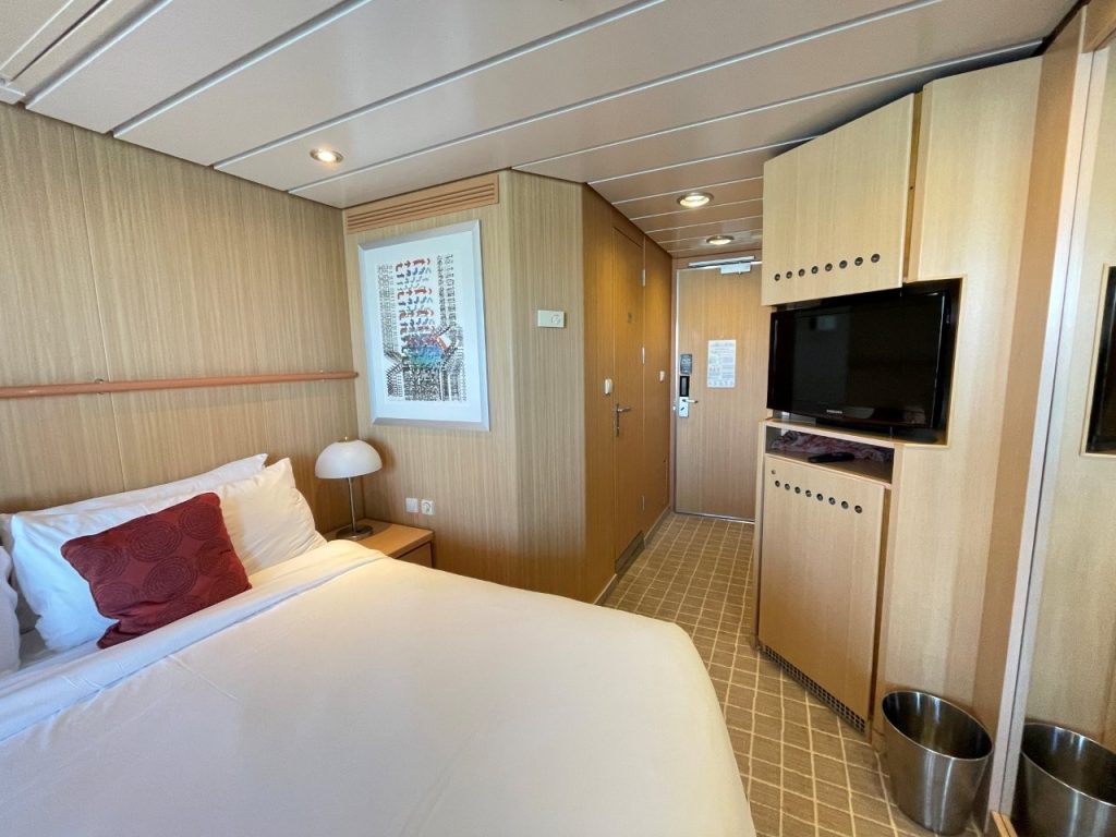 A compact Celebrity Infinity veranda cabin has a large white bed, red accent pillow, light-wood cabinetry, television and a narrow walkway leading to the door. This view shows the cabin’s somewhat dated styling and limited floor space discussed in the Celebrity Infinity review.