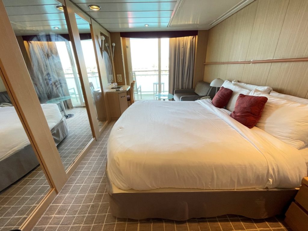 A large white bed fills much of the veranda cabin, with mirrored closet doors, a small sitting area and bright balcony doors visible beyond it. Despite its compact size, the cabin included enough seating and storage to make an eight-night Celebrity Infinity cruise comfortable.